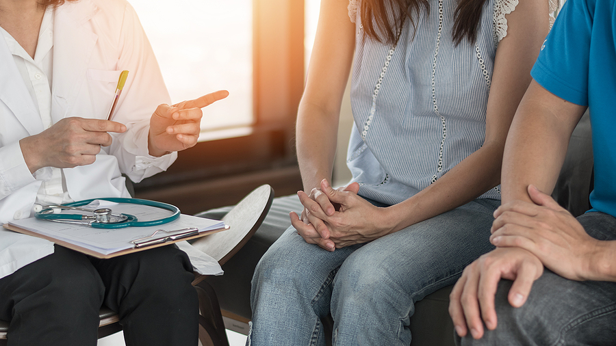 A woman and a man sit on a bench and listen to a physician talking